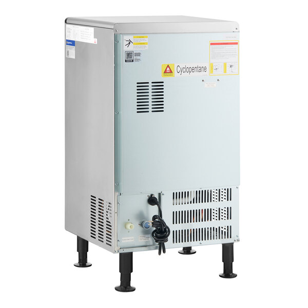 A metal undercounter ice machine with vented panels, a power cord, and warning labels, standing on four adjustable legs.