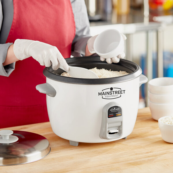 A woman in a red apron using a Main Street Equipment stainless steel lid to cook rice in a white rice cooker.