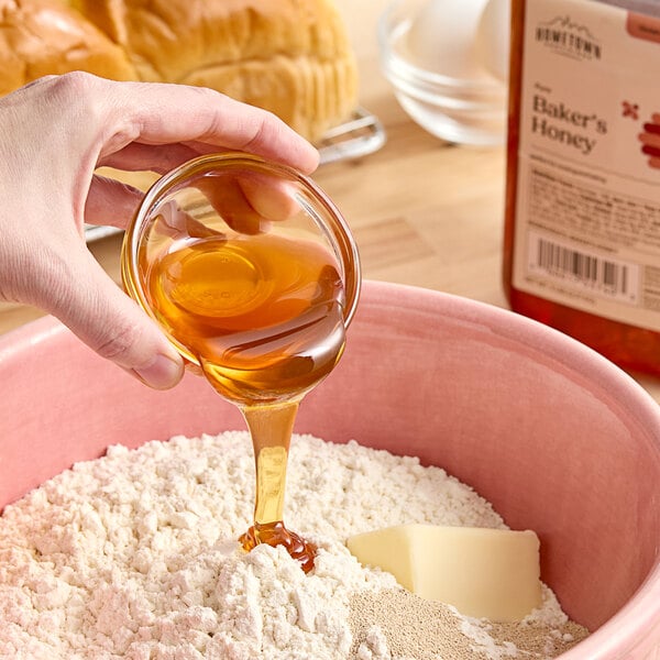 A person pouring Hometown Provisions Baker's Special honey into a bowl of flour on a counter in a home kitchen.