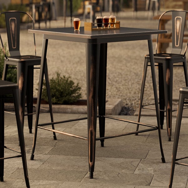 A Lancaster Table & Seating distressed copper bar height outdoor table with metal chairs on a stone patio.