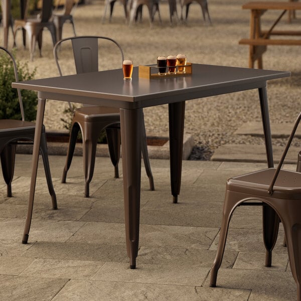 A Lancaster Table & Seating copper outdoor table on a stone patio with a brown chair under it.