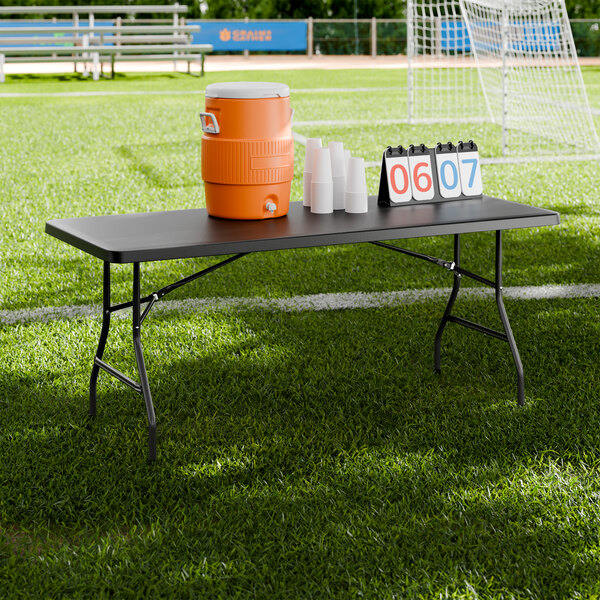 A black heavy-duty blow molded plastic folding table with black legs set up on a sports field.