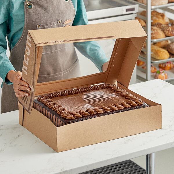 A woman holding an Innopak To Go Fresh Kraft bakery box filled with cake in front of a bakery display.