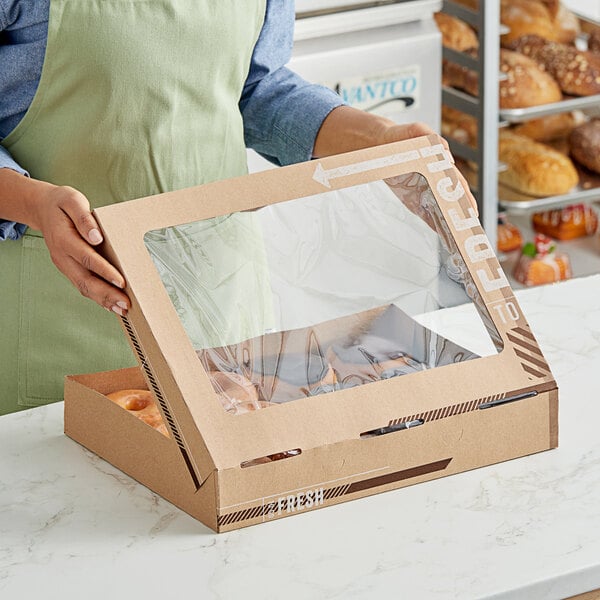 A woman holding an Innopak To Go Fresh Kraft bakery box full of donuts in front of a bakery display.