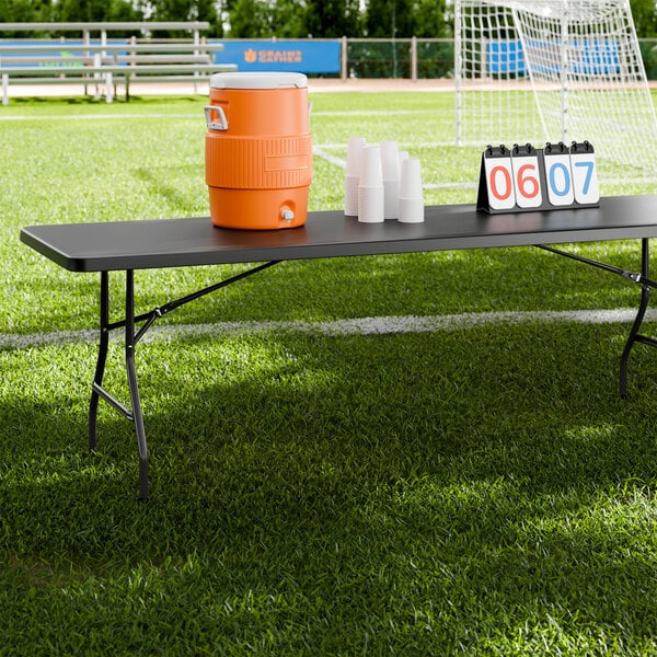 A long black heavy-duty plastic folding table with black legs set up on a sports field, holding a water cooler, plastic cups, and a scoreboard.