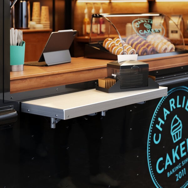 A Regency aluminum folding concession shelf mounted on a wall above a counter with a tray of pastries.