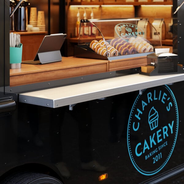 A Regency aluminum folding concession shelf mounted on a wall above a food truck with a sign that says "Charlie's Bakery"