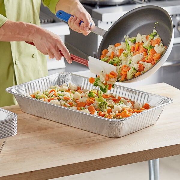 A person pouring broccoli and carrots from a tray into a Crystalware foil steam table pan.