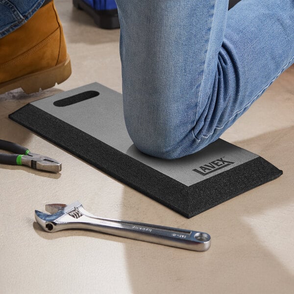 A rectangular black industrial foam kneeling pad with a handle cutout, shown being used by a person kneeling on it.