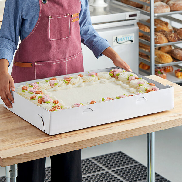 A woman wearing an apron holds a white rectangular tray of frosted cake.