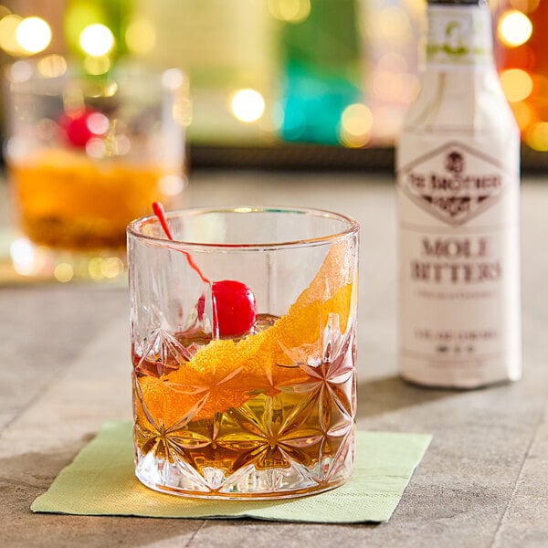 A glass of whiskey with a cherry and a straw next to a bottle of Fee Brothers Mole Bitters.