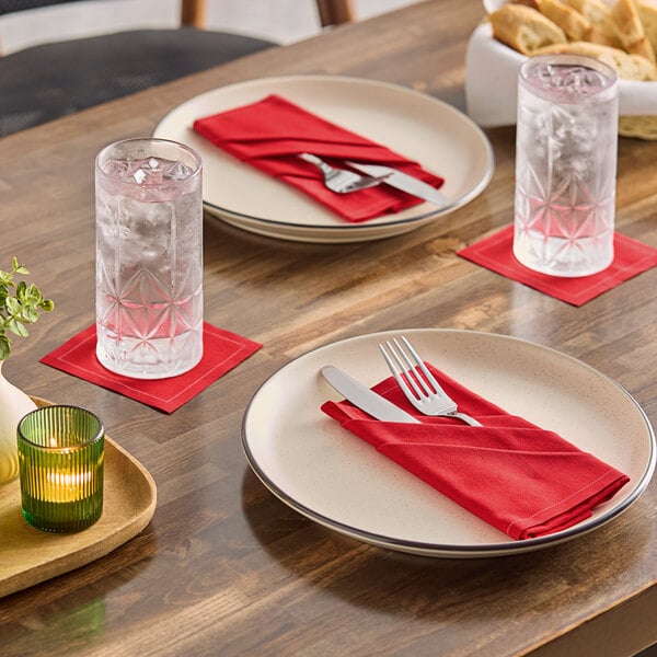 A set of beige dinner plates with red single-use cotton napkins, each holding a fork and knife, on a wooden table with glasses of water and red coasters.