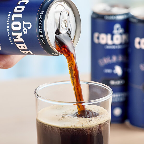 A person pouring La Colombe Black Cold Brew Coffee from a blue can into a glass of brown liquid.