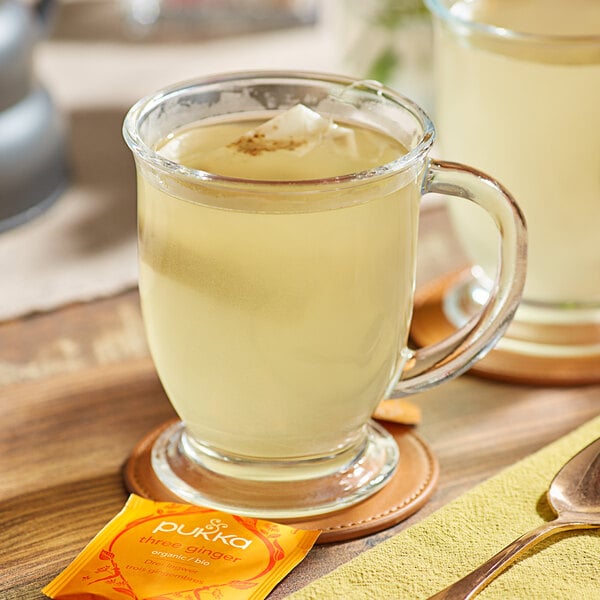 A glass mug of Pukka Organic Three Ginger tea with a tea bag on a wooden table with a spoon.