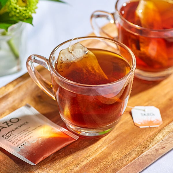 A wooden tray with two glass cups of Tazo Pumpkin Spice Chai tea with tea bags.