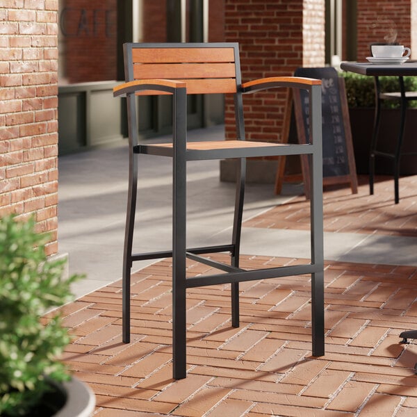 A Lancaster Table & Seating Axel series outdoor barstool with brown faux teak slats and arms on a brick patio.