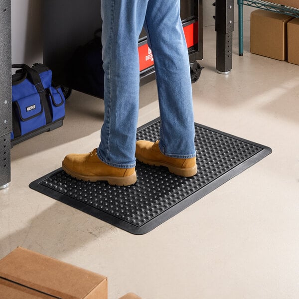 A black 2' x 3' bubble rubber anti-fatigue and anti-slip mat with a beveled edge, shown in use on a workshop floor.