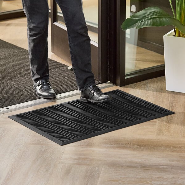 A black 2' x 3' rubber scraper mat with a wave pattern, drainage holes, and beveled edge placed at an entryway.
