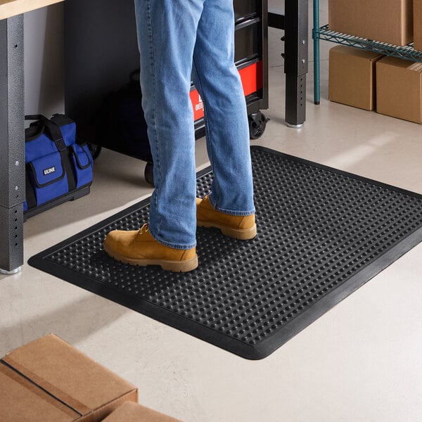 A black rubber anti-fatigue mat with a bubble surface and beveled edge placed on a workshop floor.