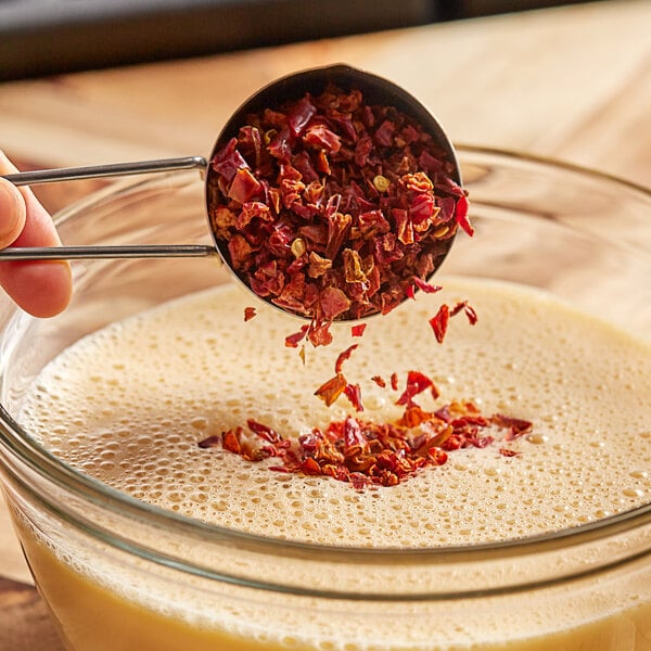 A measuring cup pouring dried red bell pepper into a glass bowl.