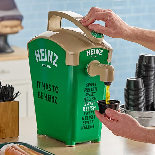A hand pouring Heinz Sweet Relish from a green and tan Heinz countertop pump dispenser.