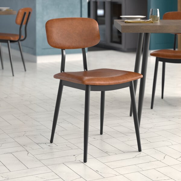 A Lancaster Table & Seating mid-century black chair with a cognac vinyl padded seat and backrest next to a table in a restaurant.