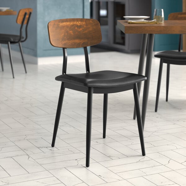 A Lancaster Table & Seating mid-century wooden chair with a black vinyl seat and wood backrest at a restaurant table.