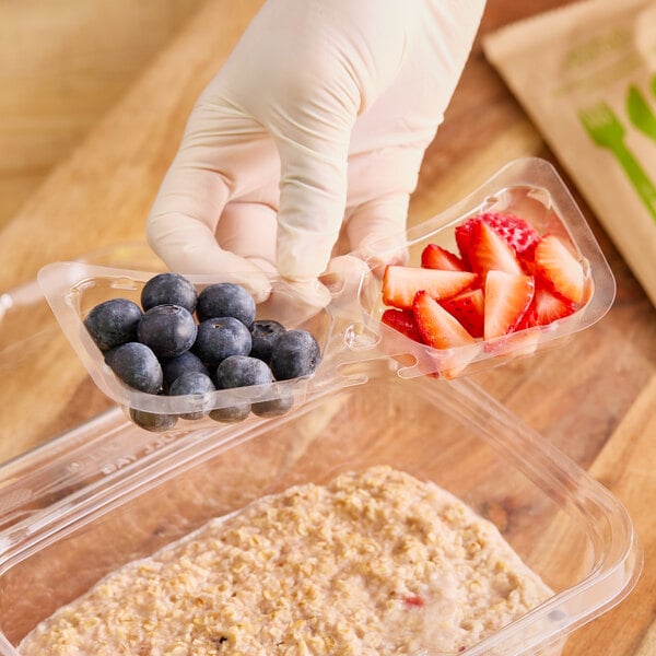 A gloved hand holding an Inline Plastics 2-compartment plastic container filled with strawberries and blueberries.