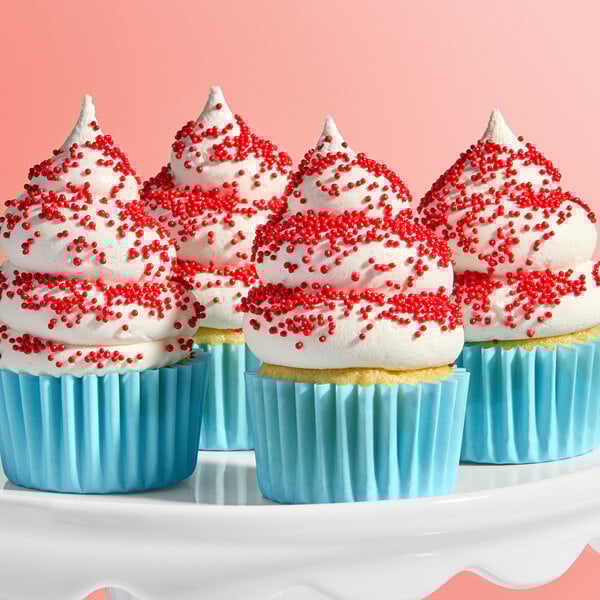 A group of cupcakes with white frosting and red nonpareil sprinkles in blue wrappers on a white cake stand.
