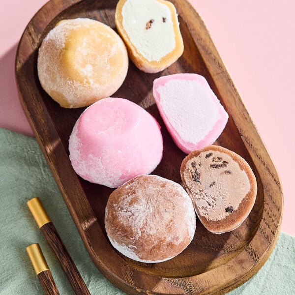 A wooden tray with assorted mochi ice cream in different flavors and colors, accompanied by a pair of chopsticks.