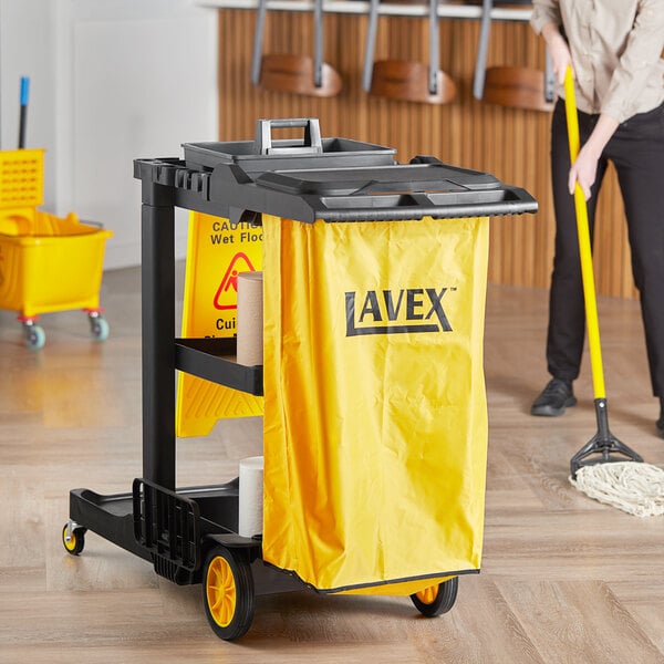 A woman cleaning a floor in a professional kitchen with a Lavex Premium janitor cart with yellow bags and a yellow mop.