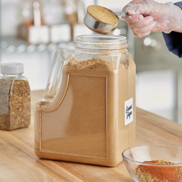 A person pouring brown powder from a metal container into a 160 oz. rectangular plastic spice container.