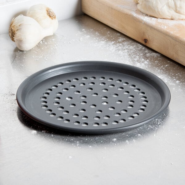 A black American Metalcraft Super Perforated Hard Coat Anodized aluminum pizza pan with holes next to garlic.