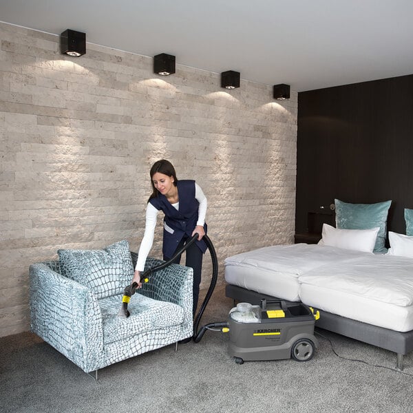 A woman using a Karcher Puzzi 10/1 C carpet spotter to vacuum a chair in a hotel room.