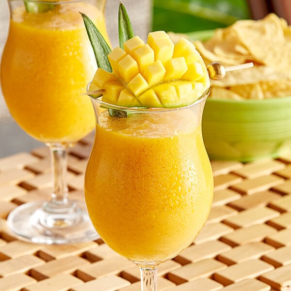 A glass of yellow mango drink with a green stem on a table.