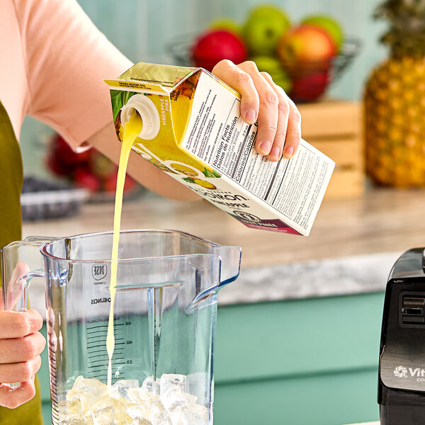 A person pouring Les Vergers Boiron Pineapple Real Fruit Puree from a 1-liter carton into a blender pitcher filled with ice.
