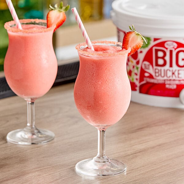 Two glasses of pink strawberry daiquiri with straws and a bottle of Master of Mixes Big Bucket Strawberry Daiquiri.