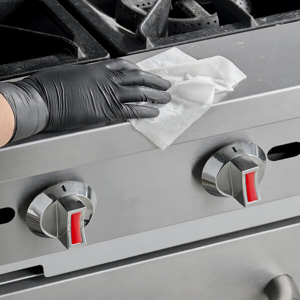 A person wearing a black glove is wiping the stainless steel surface of a stove with a white disinfecting wipe.