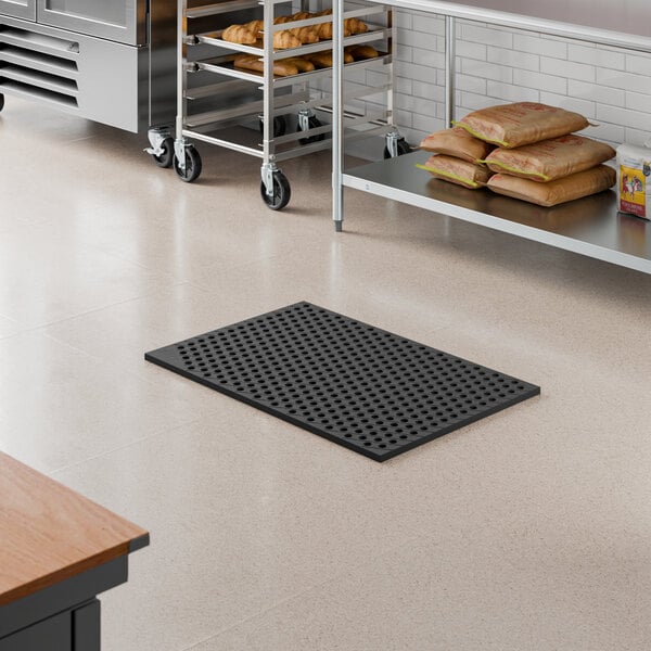 A black rubber anti-fatigue floor mat with a diamond pattern, measuring 2 feet by 3 feet and 3/4 inch thick, placed in a commercial kitchen setting.