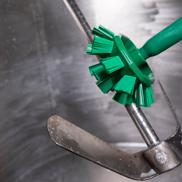 A Vikan green tank brush head with stiff polyester bristles being used to clean a metal object.