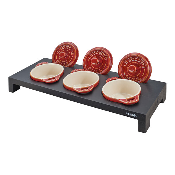 A black wooden display stand holding three red Staub mini Dutch ovens.