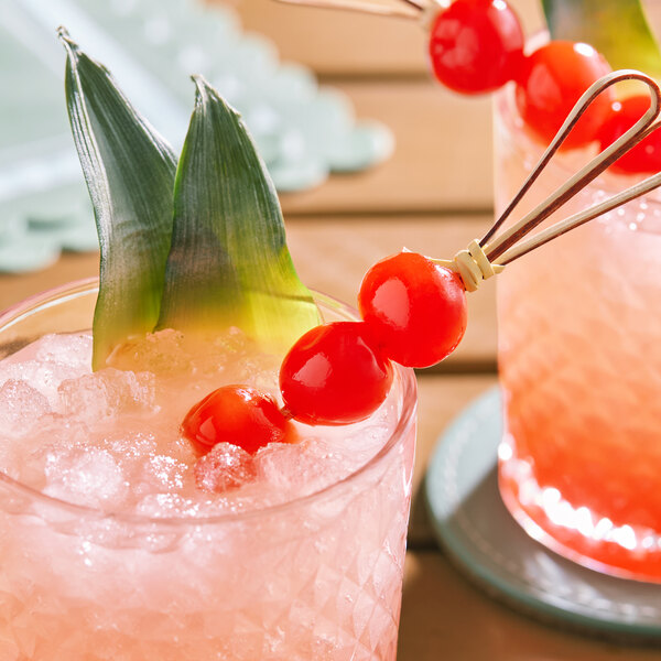 A glass of a pink cocktail garnished with maraschino cherries and pineapple leaves.