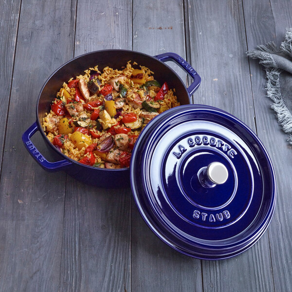 A dark blue Staub enameled cast iron dutch oven with a lid on a table with food in it.