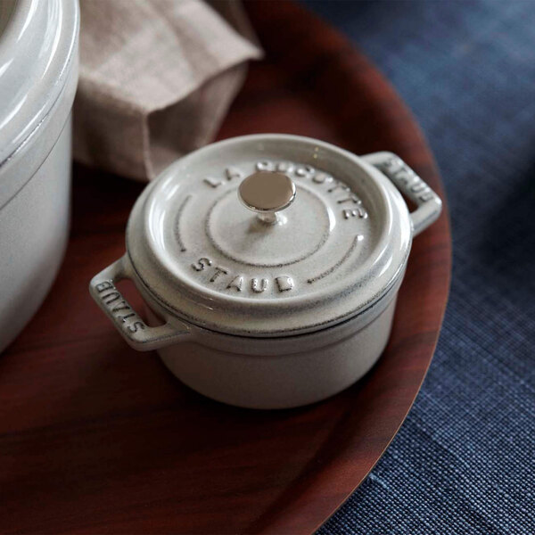 A white Staub mini enameled cast iron dutch oven with a lid on a wooden tray.