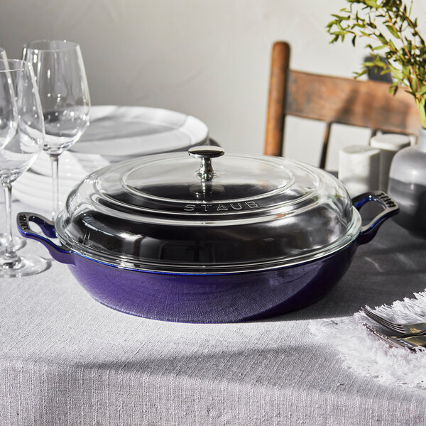 A dark blue Staub enameled cast iron brazier with a glass lid on a table with wine glasses and a plant.