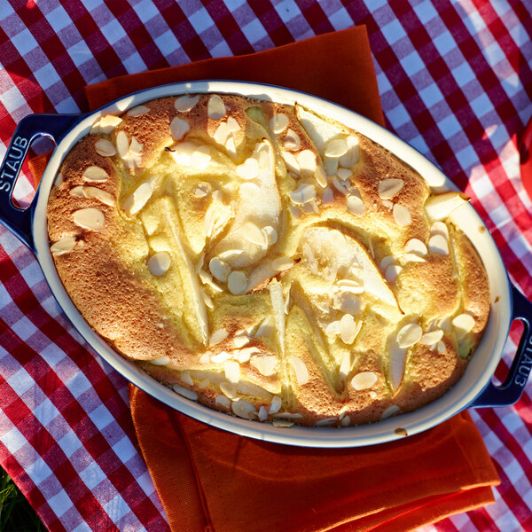 A baked dessert in a Staub dark blue ceramic oval baking dish on a table with a red and white checkered tablecloth.