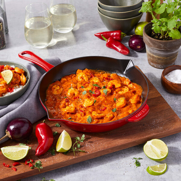 A Staub enameled cast iron skillet with shrimp and vegetables on a table.