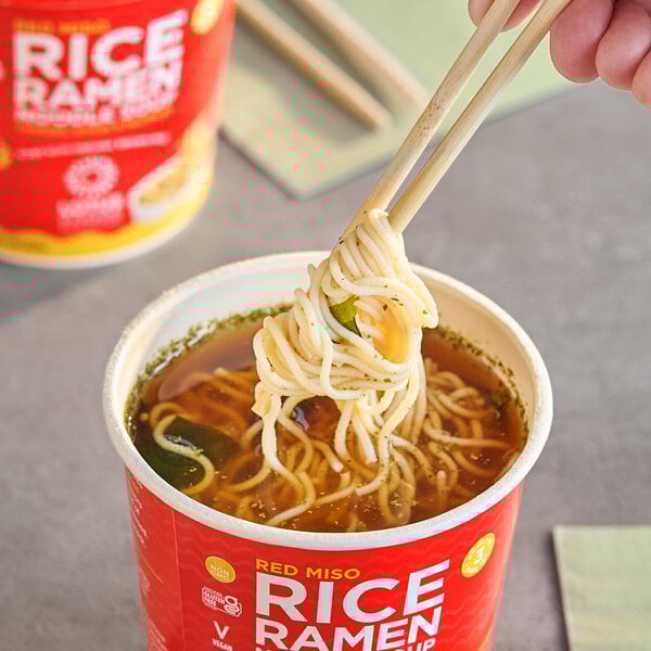 A hand holding chopsticks over a cup of Lotus Foods Red Miso Rice Ramen.