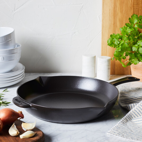 A black Staub enameled cast iron skillet on a marble counter with garlic and onions.