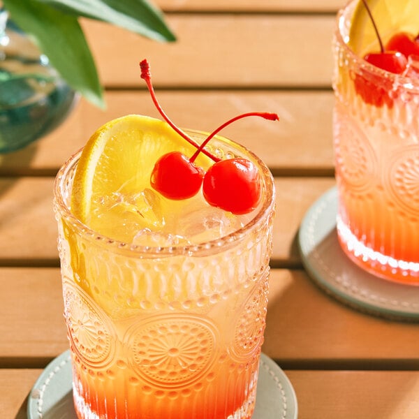 A glass of iced cocktail garnished with two maraschino cherries with stems and a lemon wedge.